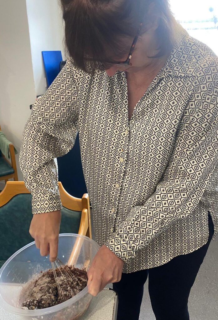 A woman in a patterned shirt is using a whisk to mix chocolate cake batter in a large plastic bowl on a table.