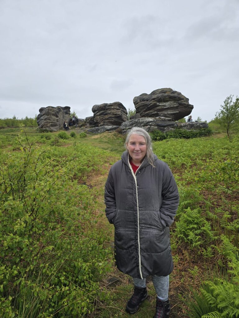 A woman stands smiling in front of the path leading toward the distinctive rock formations at the top of the hill, surrounded by green shrubs and ferns.