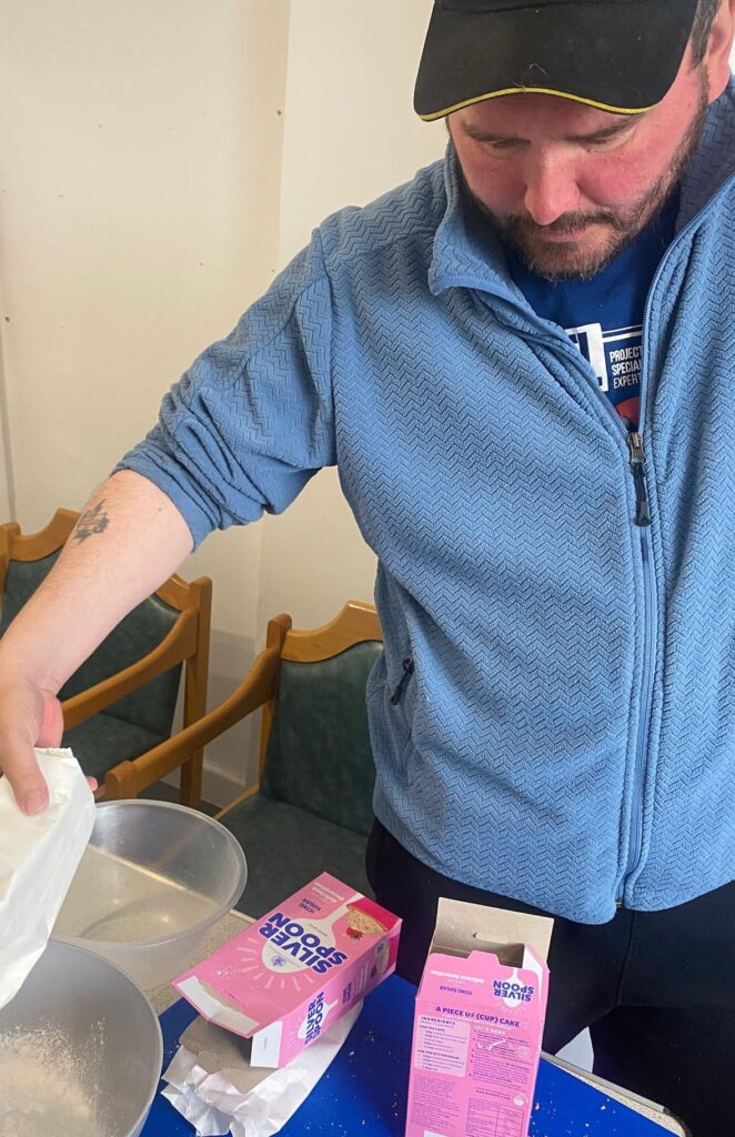 A man in a blue fleece and black cap is pouring flour into a mixing bowl. Sugar packets and other baking ingredients are visible nearby.