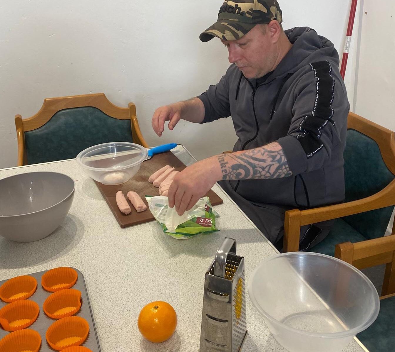 A man in a grey hoodie and camouflage cap prepares sausages at a table. A grater, orange, and silicone cupcake cases are laid out beside him.