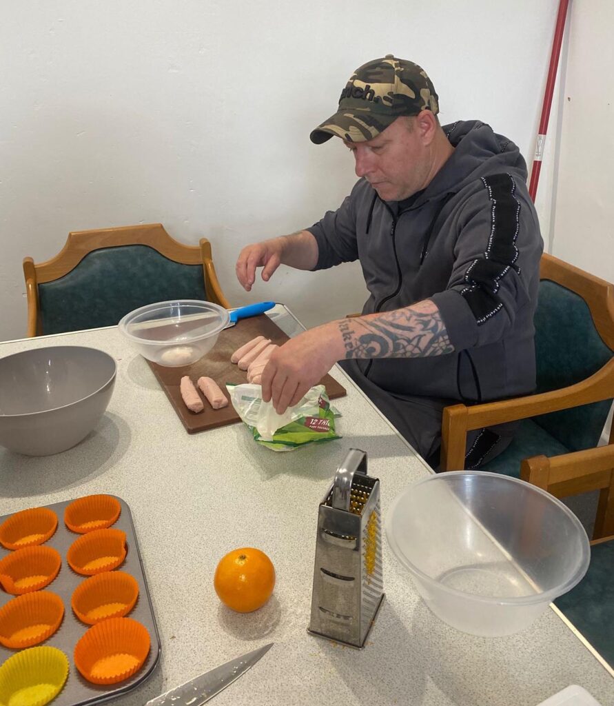 A man in a grey hoodie and camouflage cap prepares sausages at a table. A grater, orange, and silicone cupcake cases are laid out beside him.