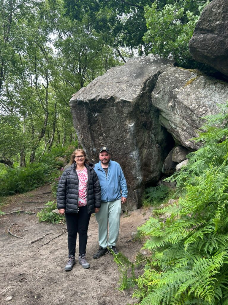 Two walkers stand smiling in front of a large boulder surrounded by lush green woodland, wearing outdoor clothing on a dirt trail.