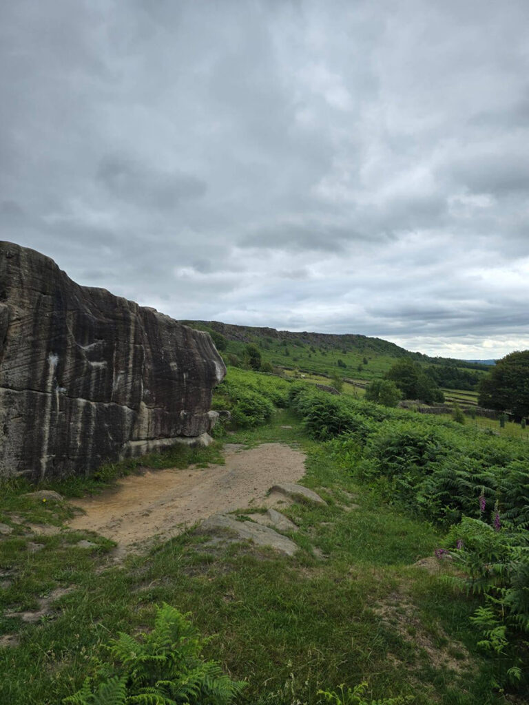 A wide path runs alongside a weathered stone outcrop, leading into green countryside under an overcast sky.
