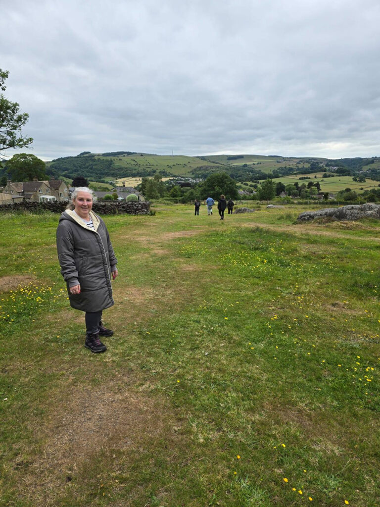 A woman in a grey coat stands on grassy open ground with scattered yellow wildflowers, with rolling hills and other walkers visible in the distance.