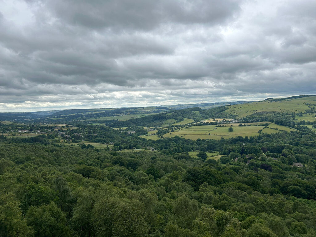 A panoramic view over the Derwent Valley shows layers of green hills and trees stretching into the distance under a cloudy sky.